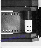 . Pull tapered end of film across back of camera and insert into and through any one of the slits of Film Take-up Spool (W).
